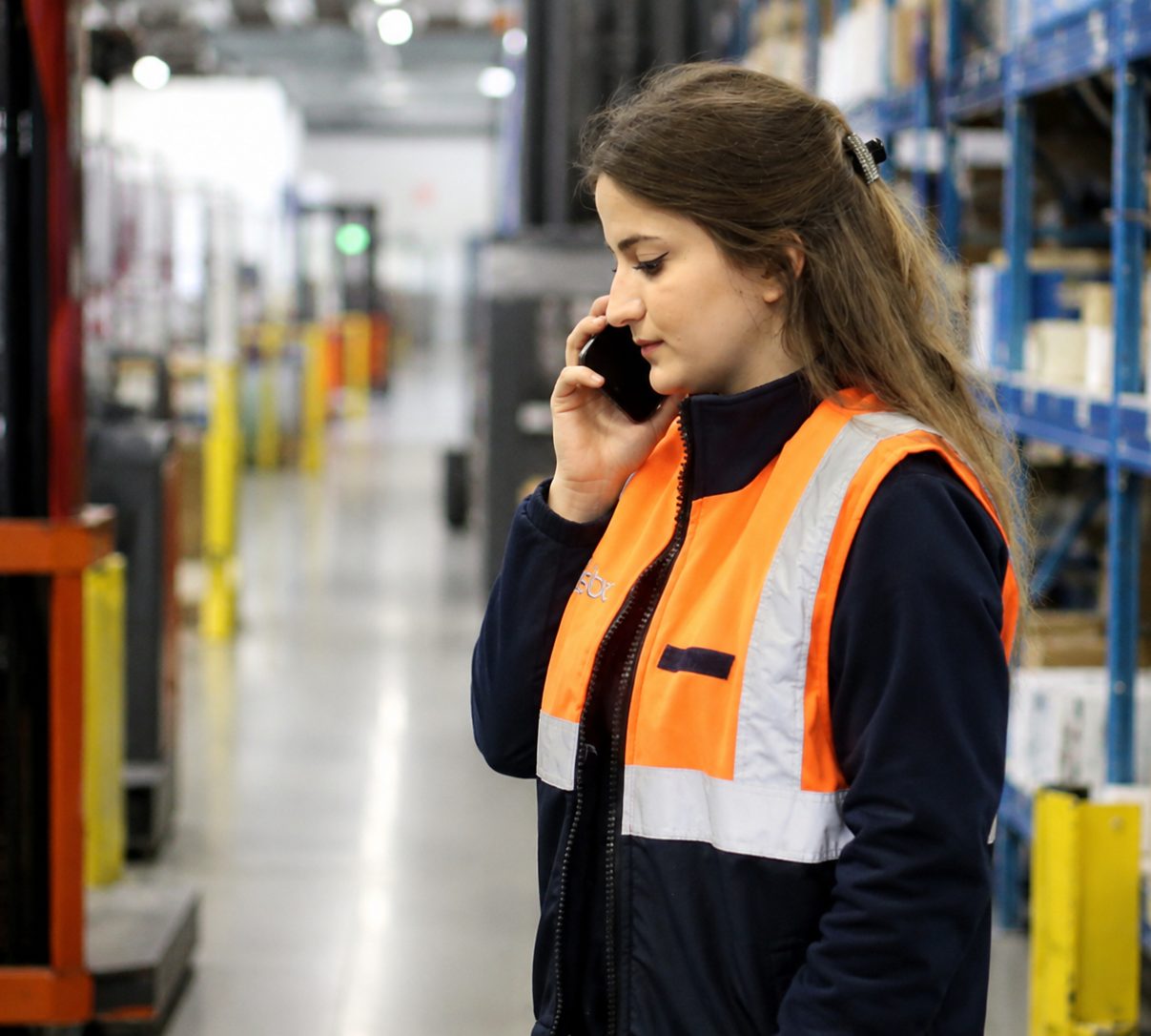 An employee in a high-visibility vest on the phone in a logistics warehouse aisle